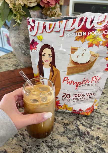 A hand holds a glass of iced coffee layered with brown liquid and cream, next to a bag of pumpkin pie-flavored protein powder, set on a speckled countertop with autumn-themed decor in the background.
