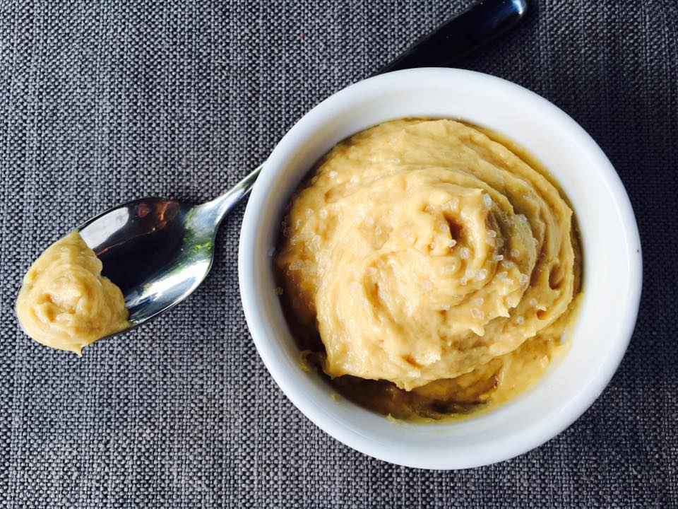 A small white bowl filled with creamy, light brown hummus sits on a textured gray surface, next to a spoon with a scoop of hummus resting on it.