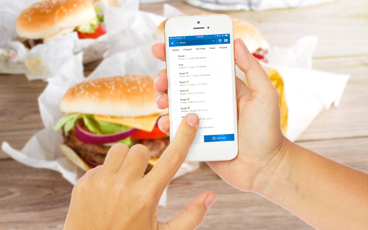 Person using a smartphone calorie-tracking app while several burgers wrapped in paper sit on a table in the background.