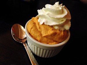 Pumpkin mousse in a white ramekin topped with whipped cream, with a spoon beside it on a dark table.