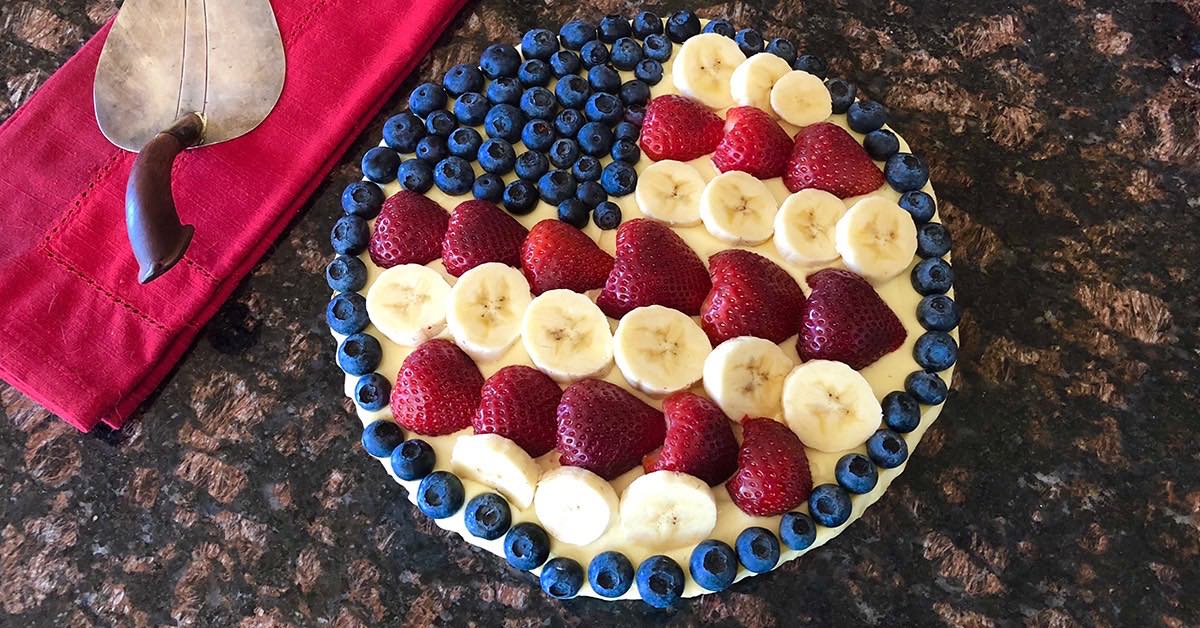 A round fruit tart decorated with strawberries, blueberries, and banana slices arranged to resemble the American flag, sits on a dark countertop beside a red cloth and a silver serving tool.