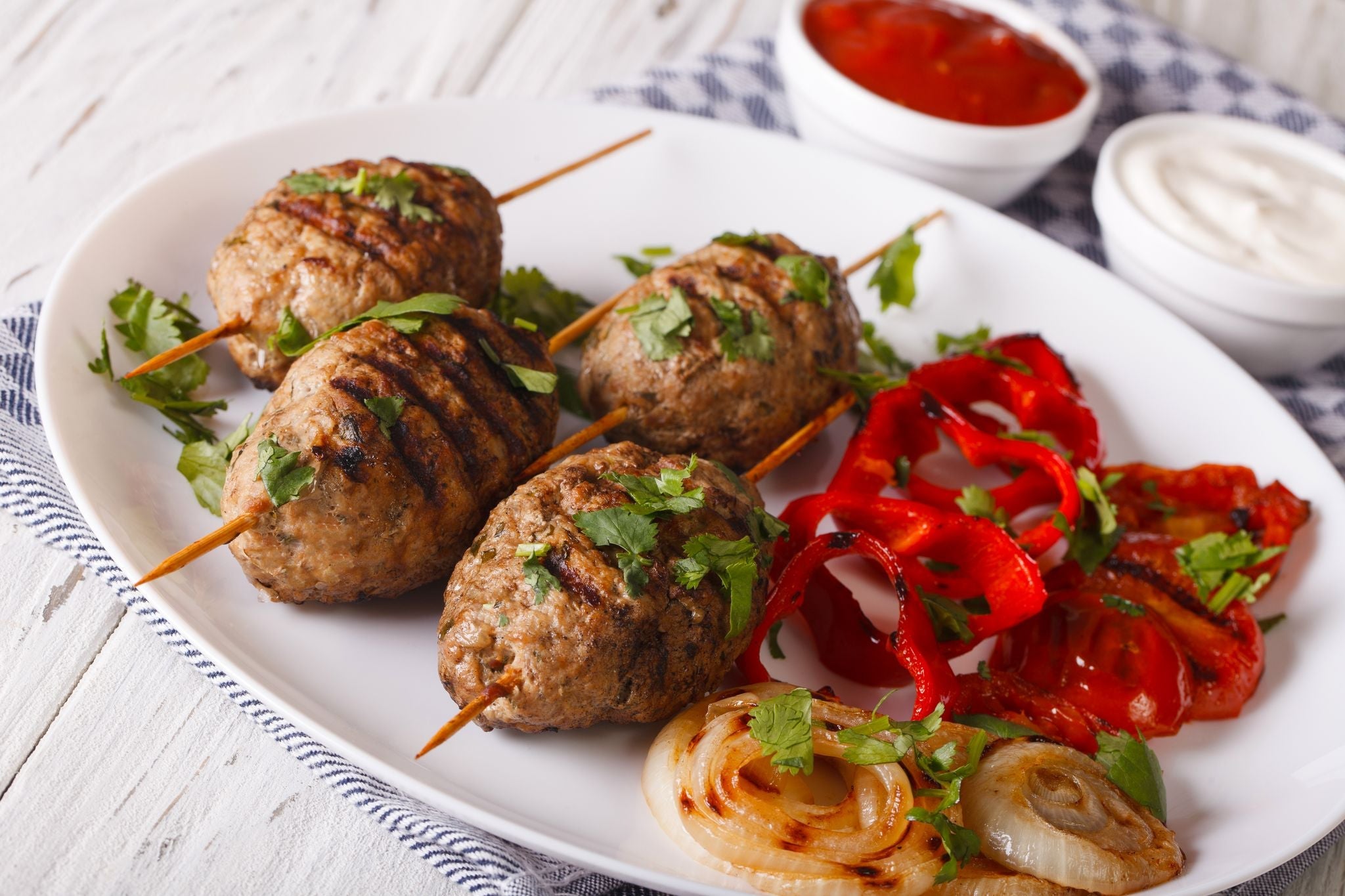 Grilled meat kebabs on skewers served with roasted red peppers, grilled onions, and fresh herbs on a white plate.