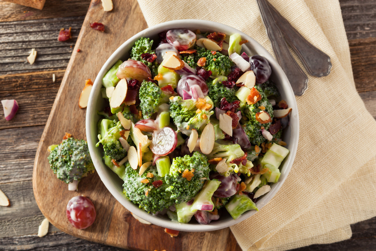 Bowl of broccoli salad with grapes, sliced almonds, bacon bits, red onion, and creamy dressing on a wooden board.