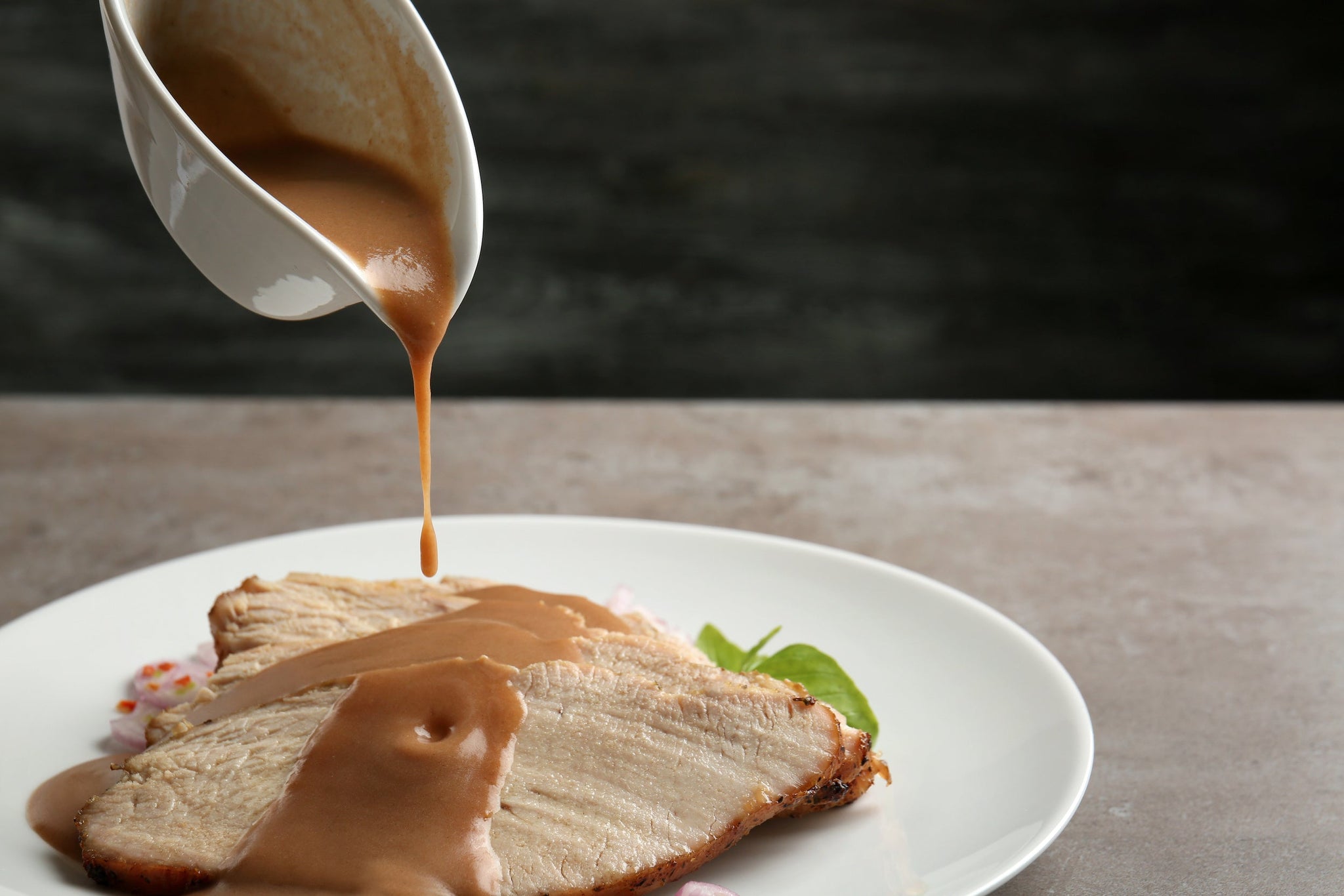 Brown gravy being poured from a white sauce boat onto sliced roasted meat on a white plate.
