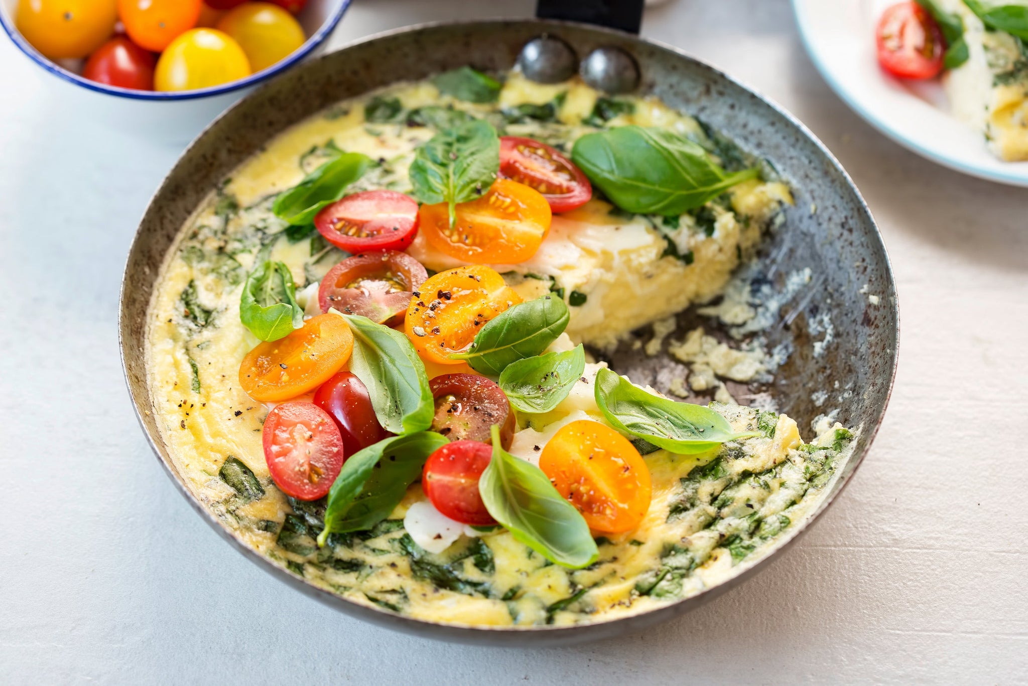 A skillet holds a fluffy frittata topped with colorful cherry tomatoes and fresh basil, with a bowl of more tomatoes in the background on a light surface.