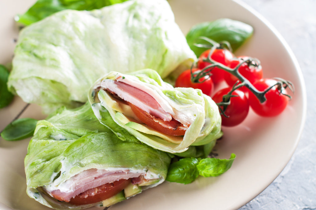Two lettuce wraps filled with ham, cheese, and tomato are placed on a beige plate, accompanied by a cluster of cherry tomatoes and fresh basil leaves.