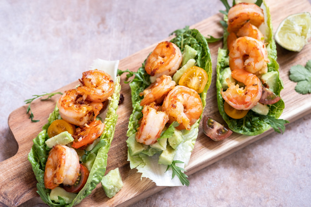 Three lettuce wraps filled with grilled shrimp, avocado, and cherry tomatoes are arranged on a wooden serving board, garnished with lime and fresh herbs.