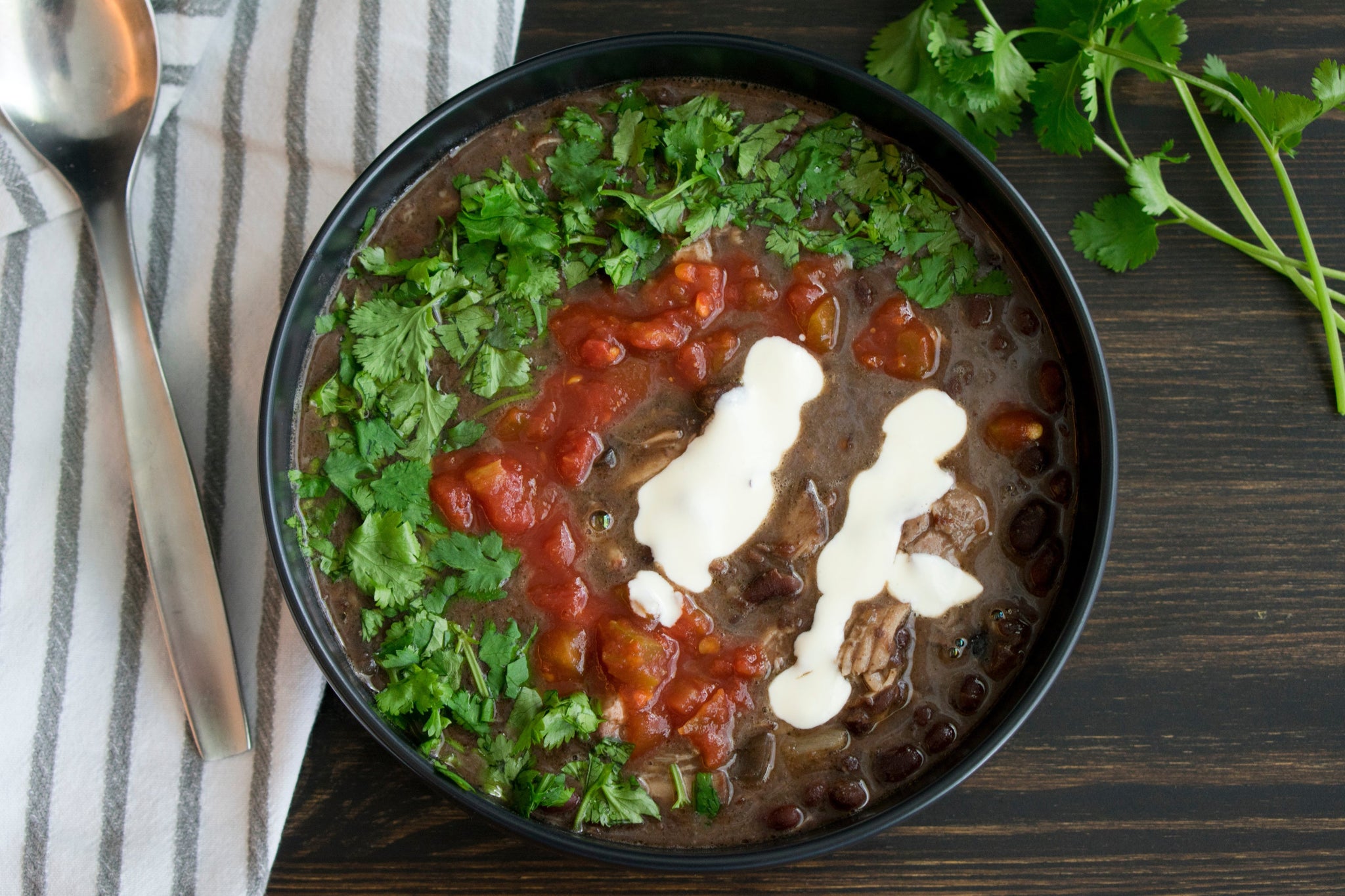 Bowl of black bean soup topped with cilantro, salsa, and sour cream, with a spoon and napkin beside it.