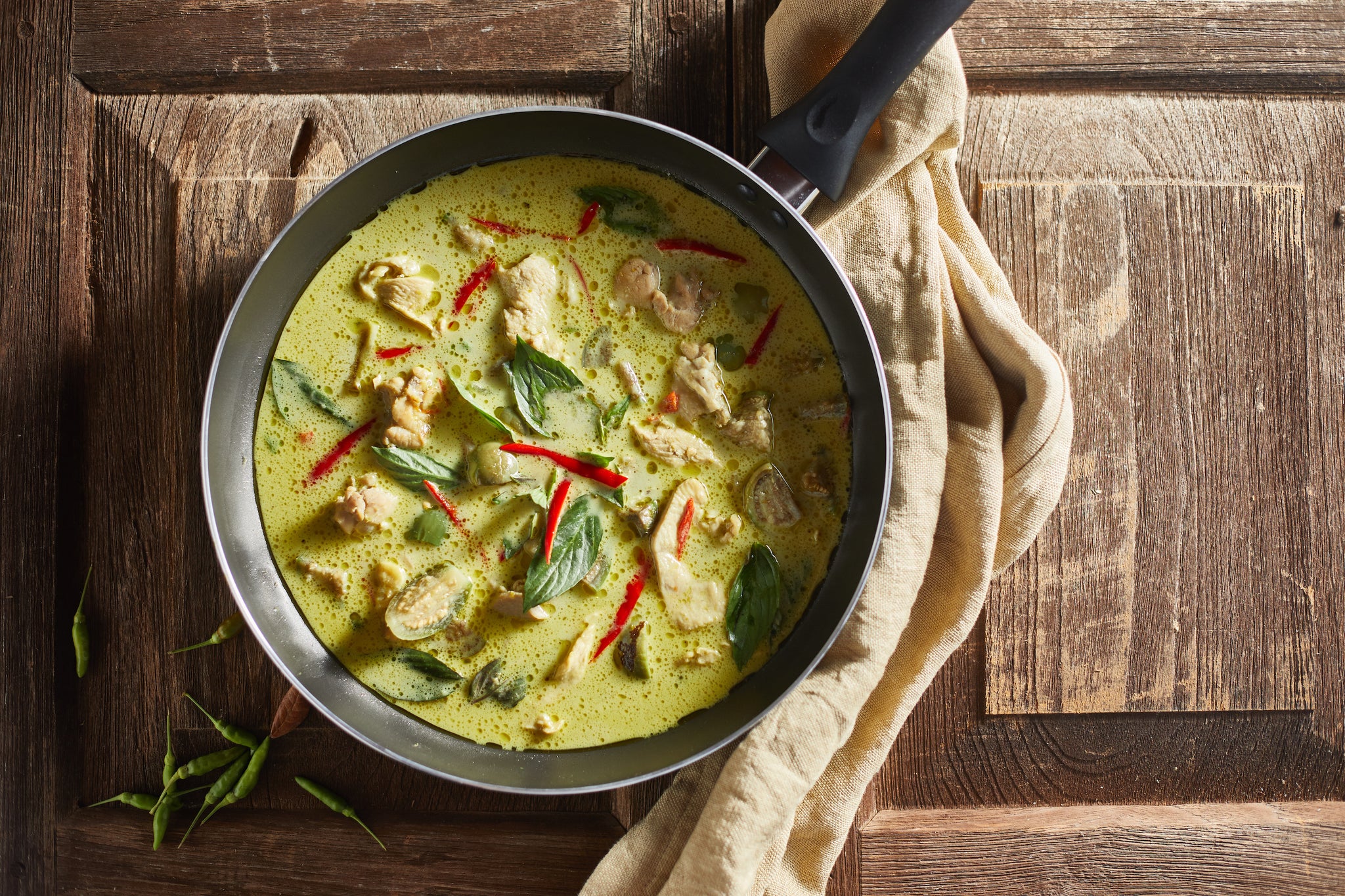 A skillet filled with green curry featuring pieces of chicken, red chili strips, and fresh basil leaves sits on a wooden surface, with a beige cloth draped beside it.