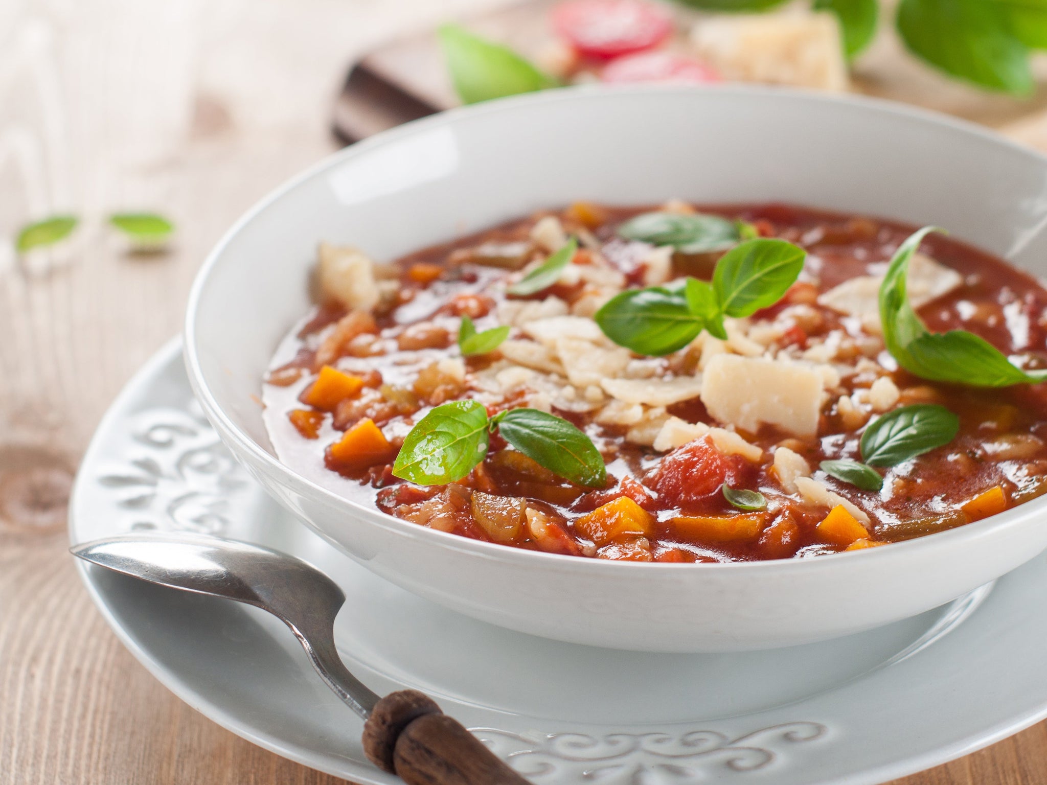 A bowl of minestrone soup filled with vegetables, topped with fresh basil leaves and grated cheese, sits on a decorative plate against a wooden background.