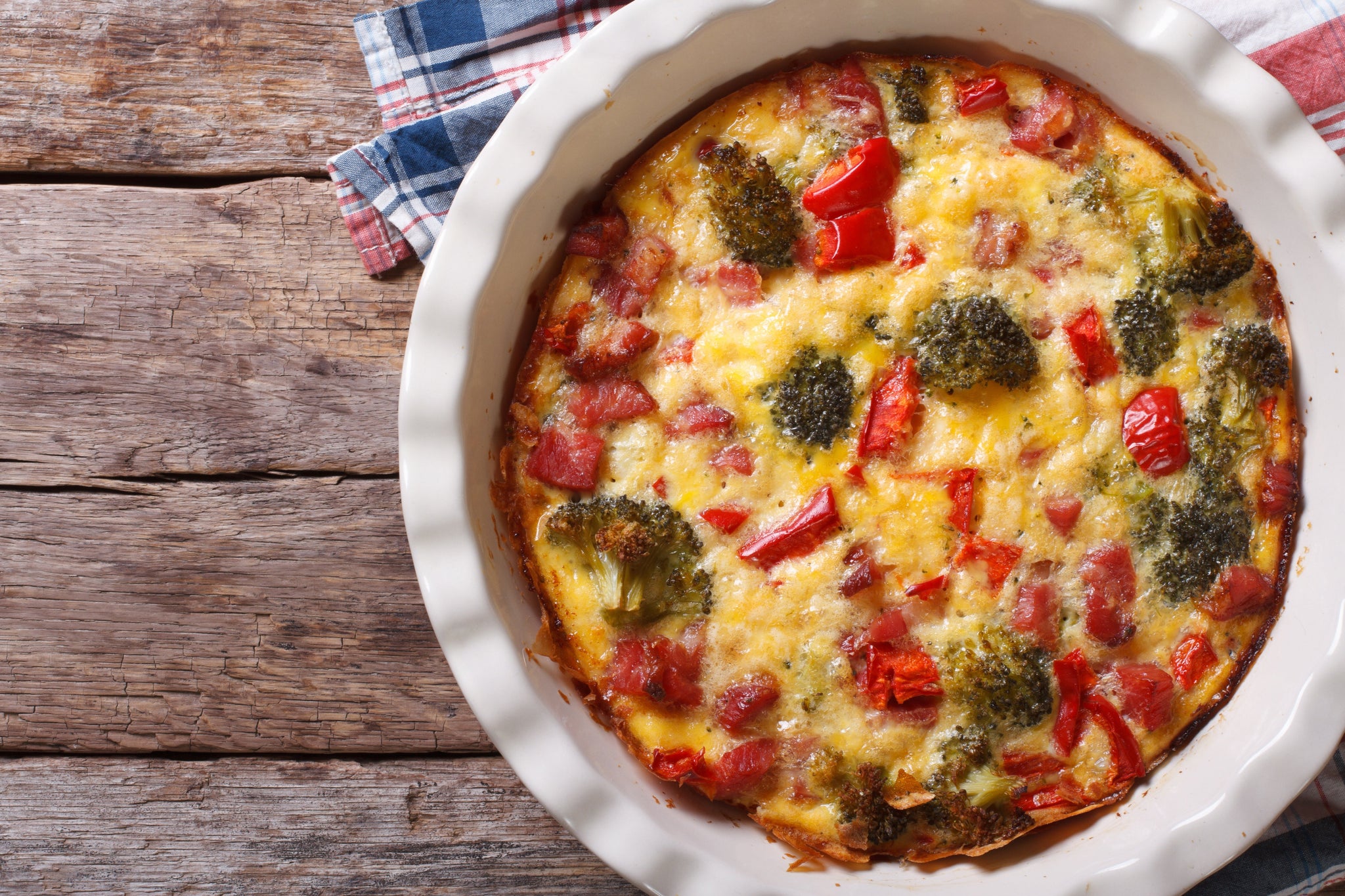 A baked vegetable and cheese frittata with broccoli and red peppers in a round white dish on a wooden table.