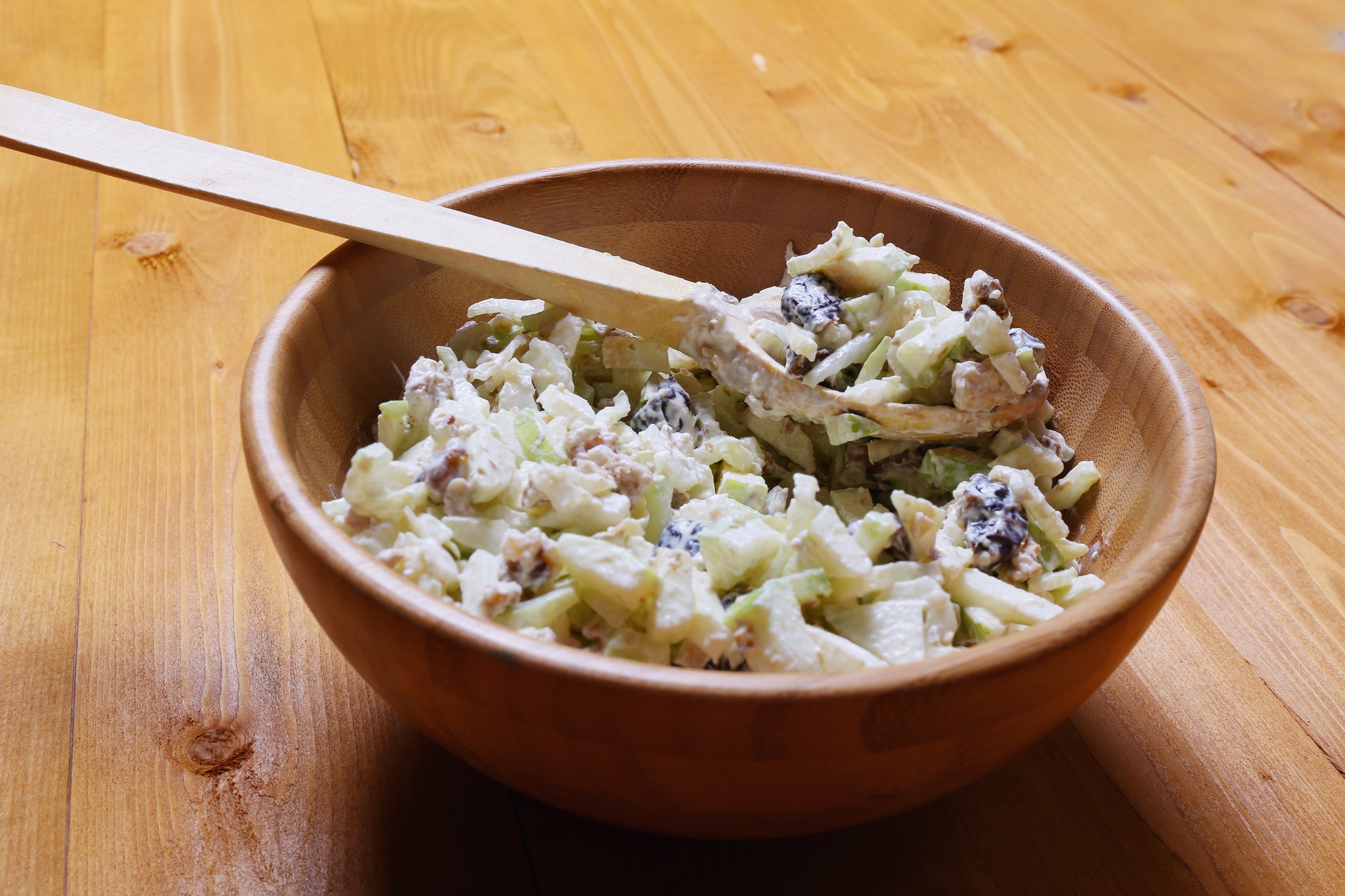 Wooden bowl filled with creamy apple salad mixed with nuts and raisins, with a wooden spoon resting inside.
