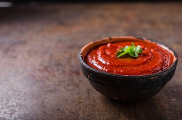 Bowl of smooth red tomato sauce garnished with a small sprig of green herb on a dark textured surface.