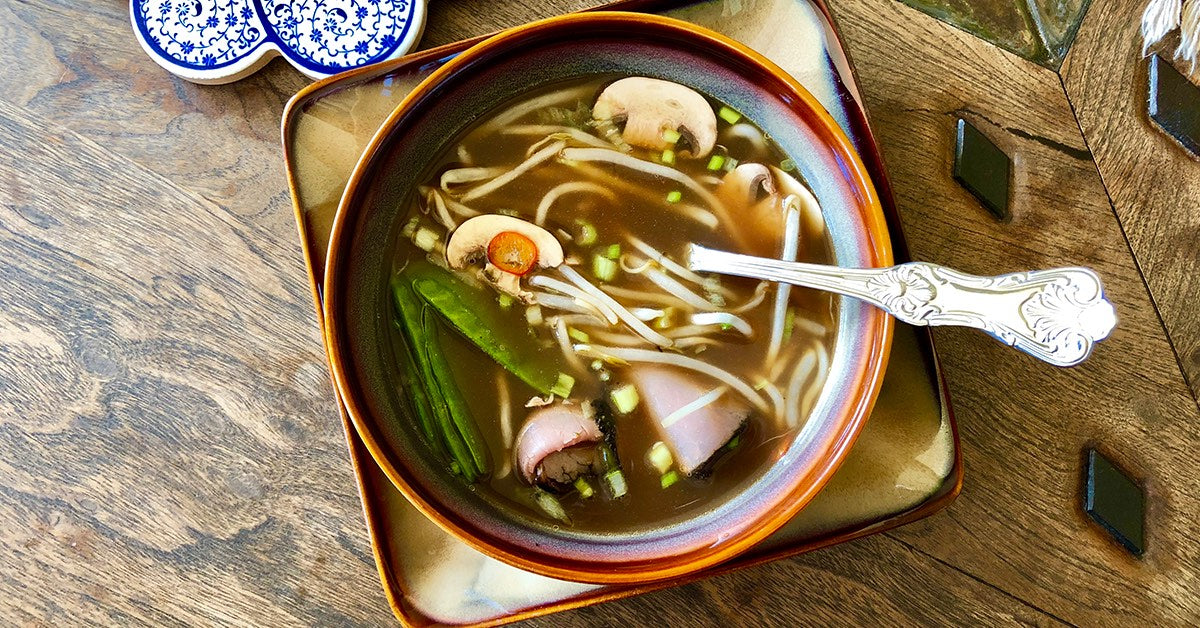 Bowl of noodle soup with mushrooms, sprouts, and snap peas on a wooden table.