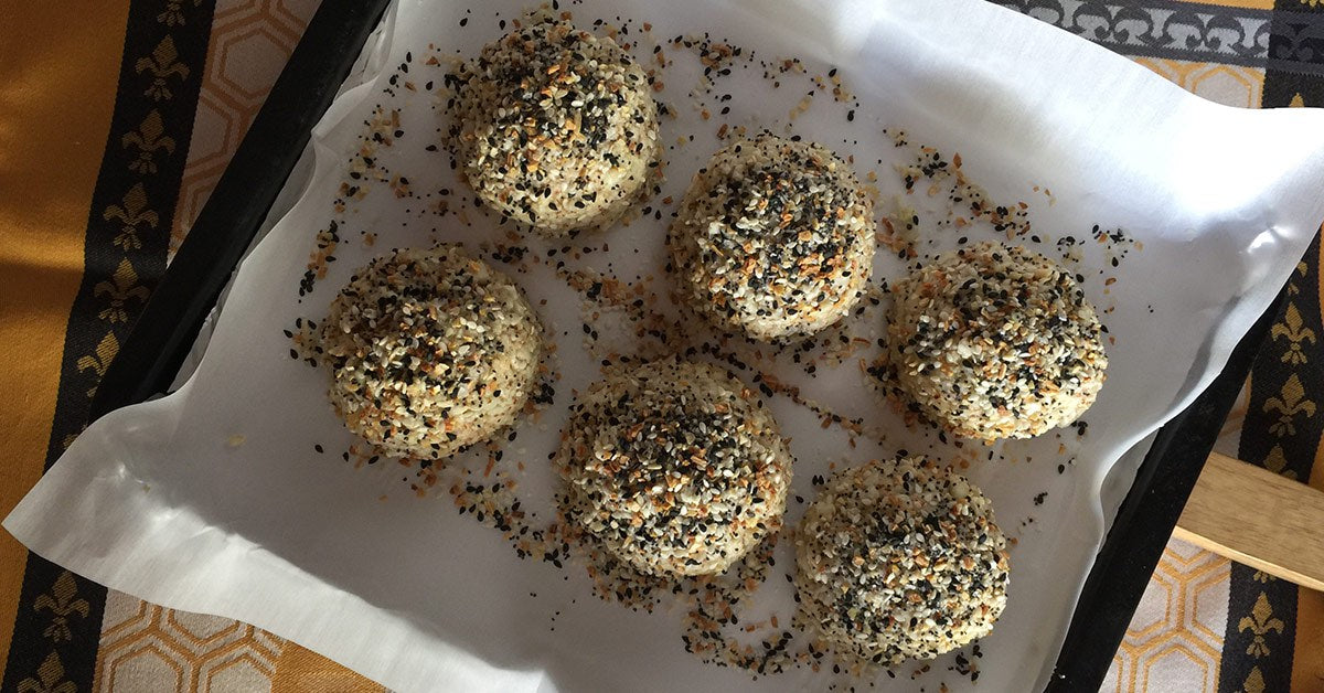 Six round, sesame-coated rice balls are arranged on parchment paper, sprinkled with various seeds, on a patterned tablecloth in warm lighting.