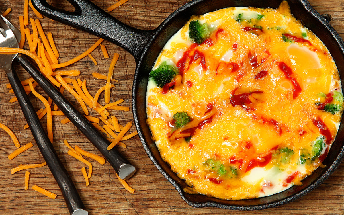 A cast iron skillet holds a cheesy broccoli and egg dish topped with ketchup, surrounded by scattered orange cheese shreds and silver utensils on a wooden surface.