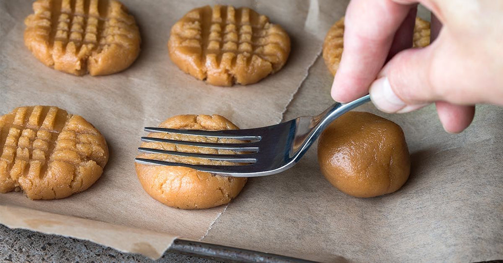 A hand presses a fork onto a ball of peanut-butter cookie dough to create a crisscross pattern on a baking sheet.