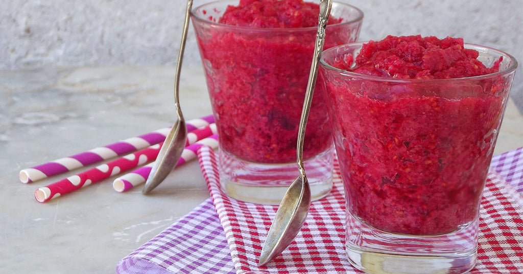 Two glasses filled with bright red berry slush, with spoons and striped straws on checkered cloths.