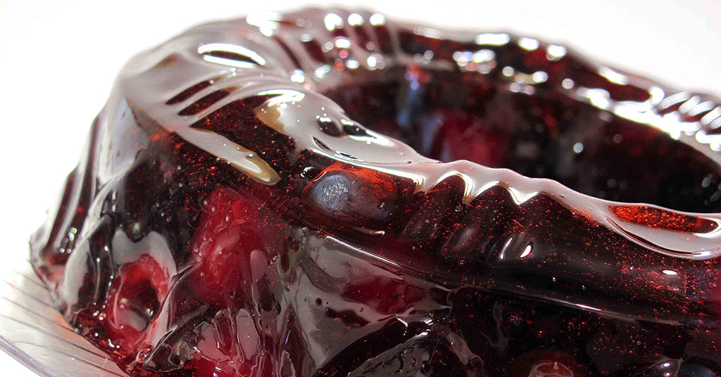 A shiny, red gelatin dessert is displayed in a decorative mold, showcasing its smooth surface and embedded fruit pieces, with light reflecting off the glossy texture.