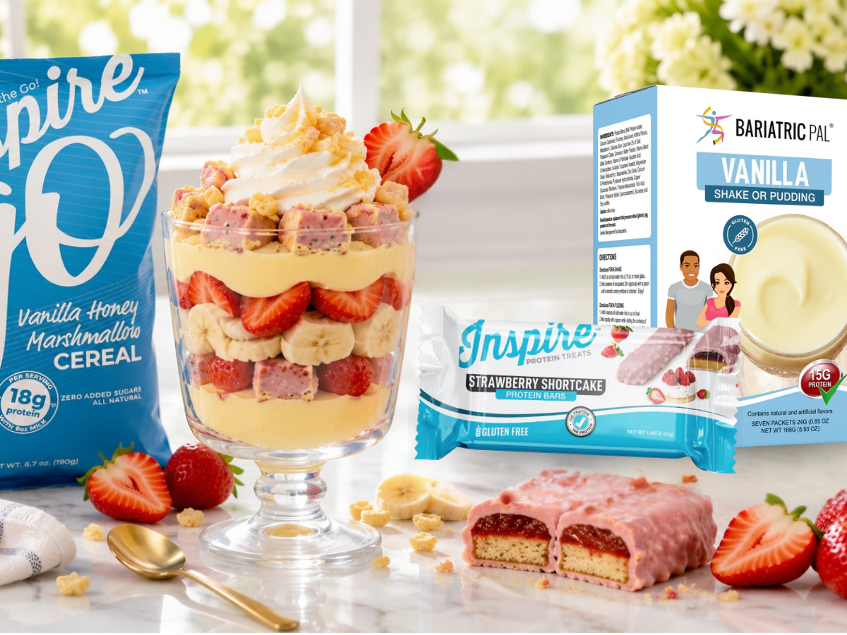 Layered strawberry shortcake protein pudding parfait with whipped cream, strawberries, bananas, cereal, and sliced protein bars, surrounded by Inspire and BariatricPal products on a bright marble countertop.