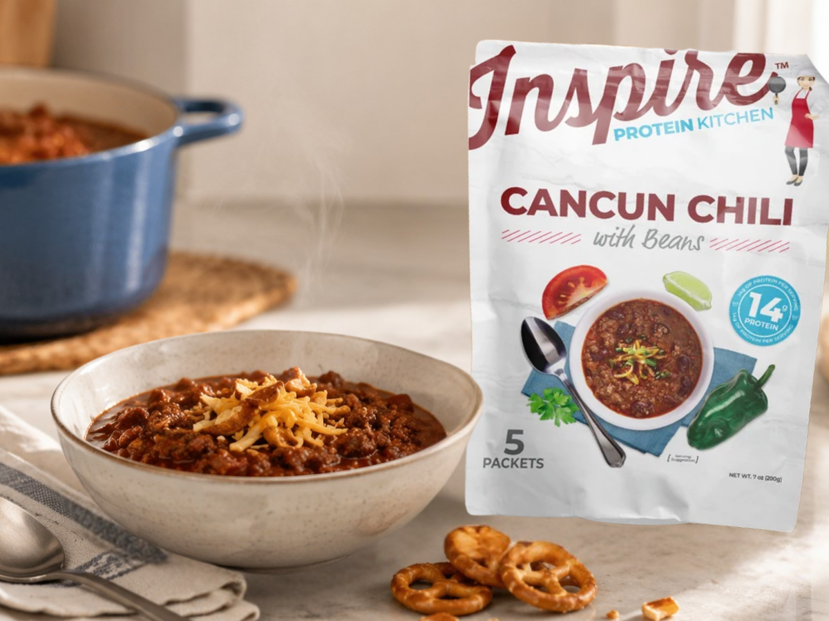 A steaming bowl of Cancun chili topped with cheese sits on a table next to a packet of Inspire Protein Kitchen chili mix and a plate of pretzels, with a blue pot in the background.