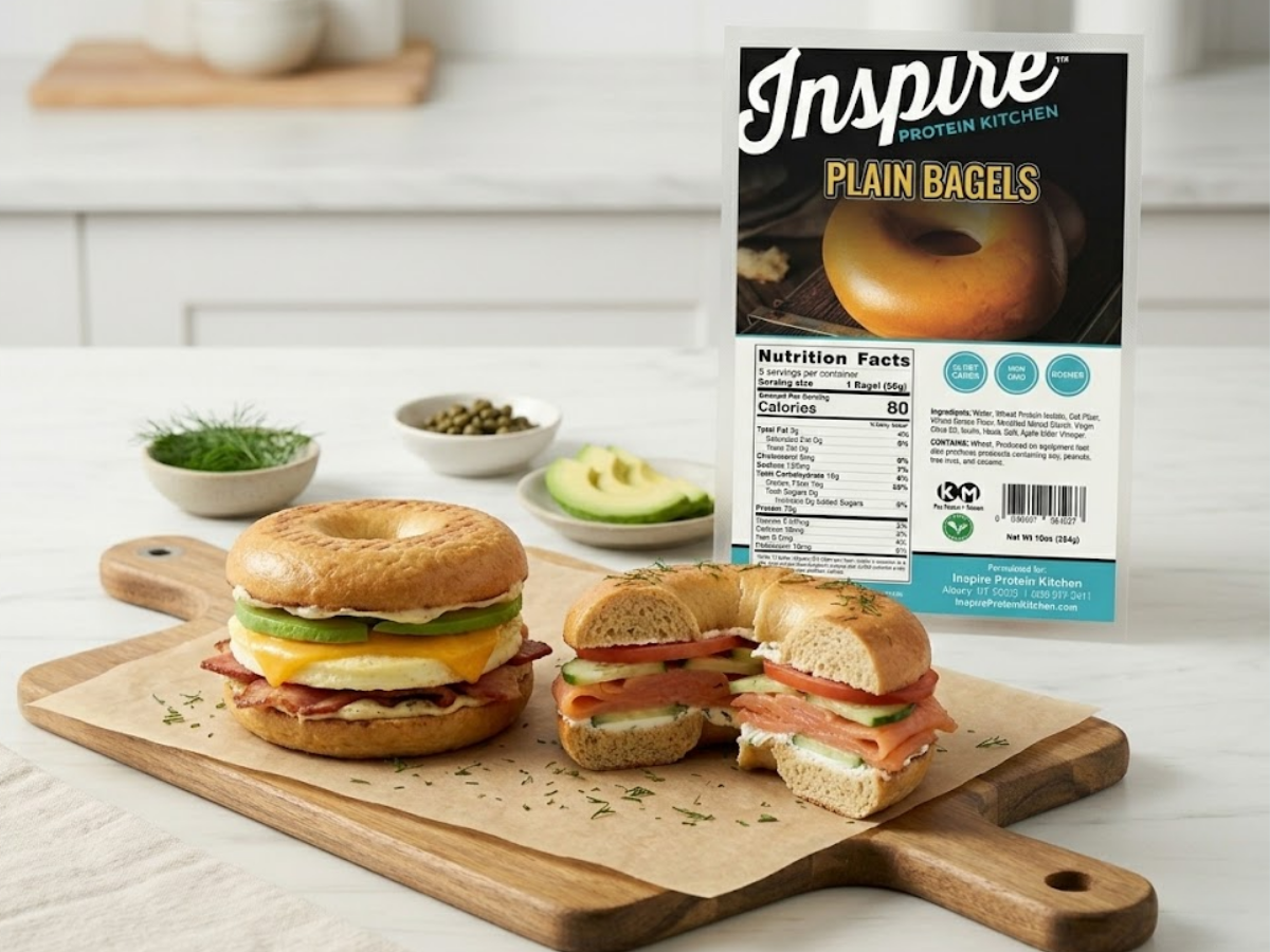 High-protein breakfast bagel sandwiches made with Inspire Protein Kitchen Bagels filled with egg, bacon, avocado, and smoked salmon, with the Inspire Protein Kitchen Bagels packaging displayed beside them in a bright kitchen setting.
