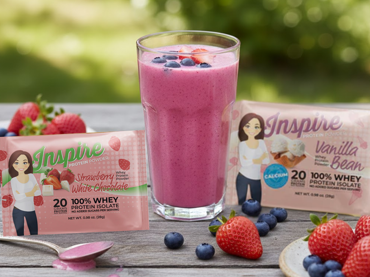 Tall glass of pink summer berry protein smoothie topped with strawberries and blueberries, placed beside Inspire Strawberry White Chocolate and Inspire Vanilla Bean protein powder packets on a wooden table outdoors.