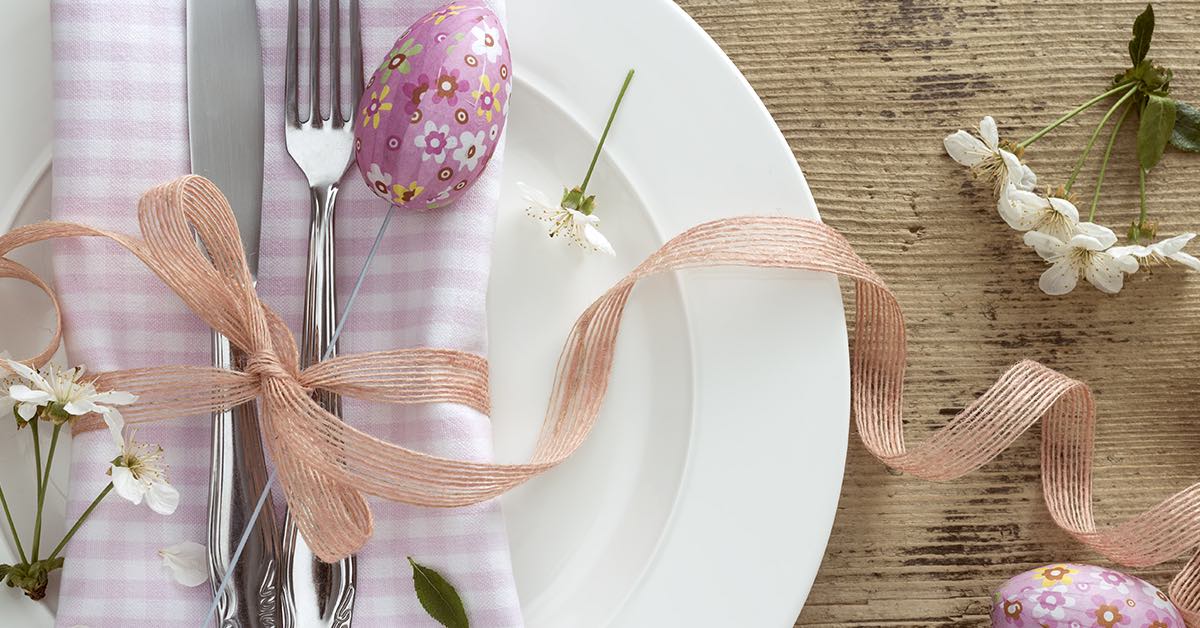 A white plate with a pink checkered napkin, silver cutlery, a decorated Easter egg, and delicate flowers is elegantly arranged with a ribbon on a rustic wooden table.