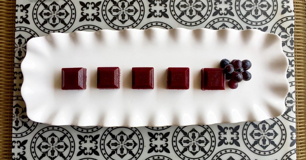 Five red square jelly candies arranged in a row on a white platter with a small cluster of grapes.
