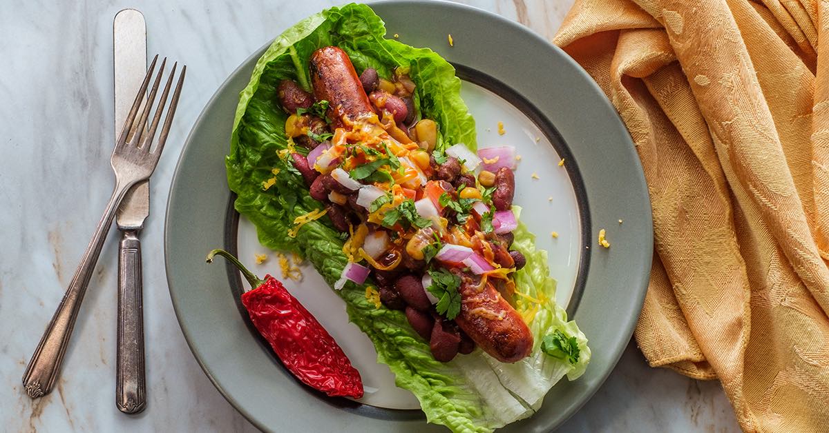 Lettuce-wrapped sausage topped with beans, onions, cheese, and herbs, served with a red chili pepper on the plate.
