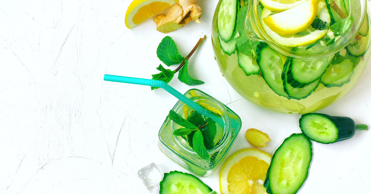 A glass pitcher filled with cucumber slices, lemon wedges, and mint leaves sits beside a small jar of mint-infused drink with a turquoise straw, all arranged on a light textured surface.