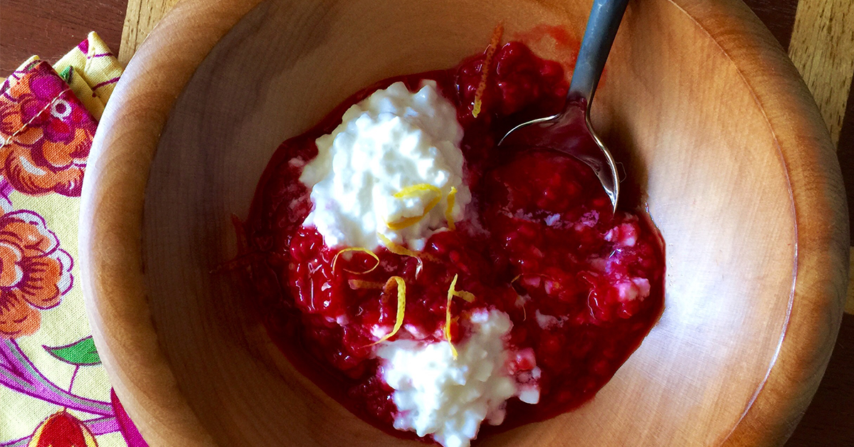 Wooden bowl with raspberry sauce and dollops of cottage cheese, topped with lemon zest and a spoon.