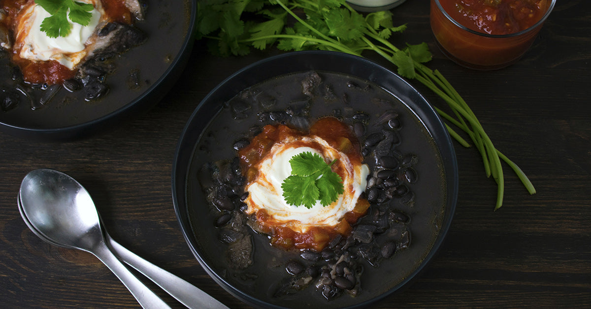 A black bean soup is served in a dark bowl, topped with sour cream, salsa, and cilantro, alongside fresh herbs and a spoon on a wooden table.