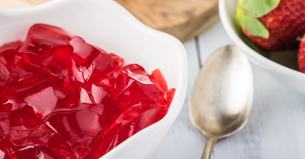 Bright red strawberry gelatin in a white bowl beside a spoon and fresh strawberries