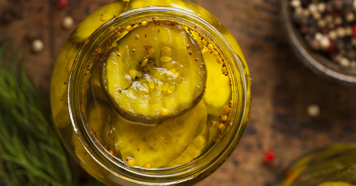 A jar filled with bright yellow bread and butter pickles sits on a wooden surface, surrounded by scattered peppercorns and fresh dill, showcasing the pickles' glossy texture and spices.