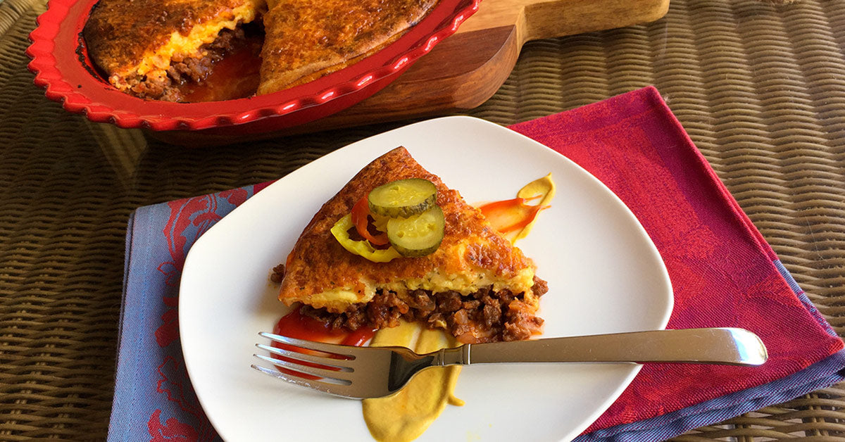 Slice of cheesy meat pie topped with pickles, served with ketchup and mustard on a white plate.