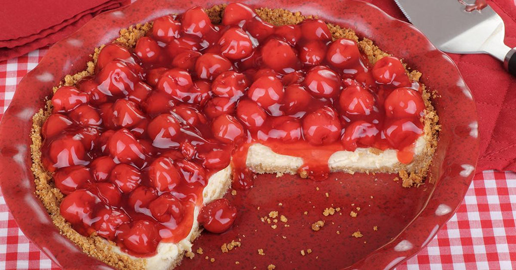 Cherry cheesecake with a graham crust, topped with glossy cherries and a slice removed.