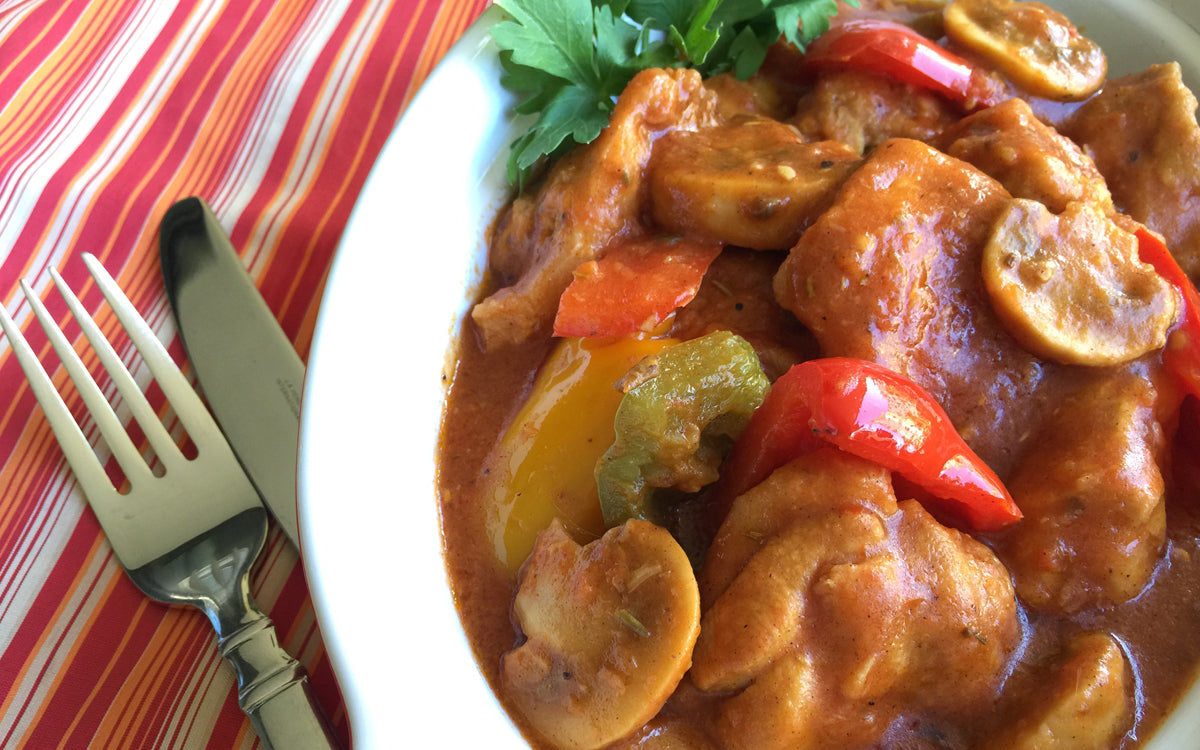 Bowl of creamy stew with chicken, mushrooms, and peppers, garnished with parsley beside a fork and knife.