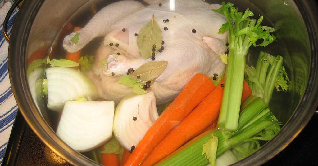 Whole raw chicken in a pot with carrots, celery, onion, bay leaves, and peppercorns ready for boiling.