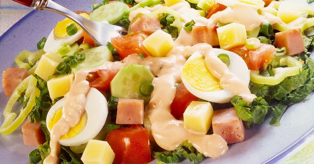 Salad with lettuce, tomatoes, cucumbers, ham cubes, cheese, sliced eggs, and creamy dressing on top.
