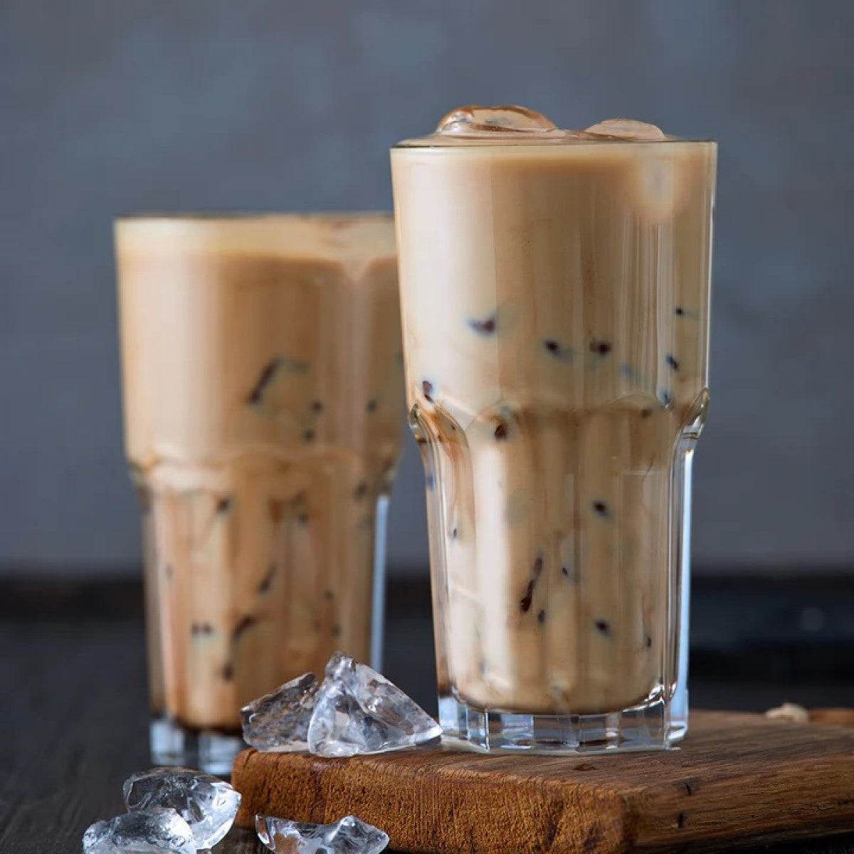 Two tall glasses of iced coffee sit on a wooden surface, filled with creamy brown liquid and ice cubes, against a soft gray background.