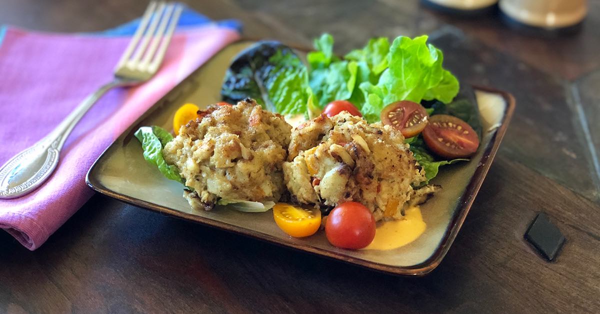 Plate with two seasoned crab cakes on greens, surrounded by cherry tomatoes, with a fork on a purple napkin.
