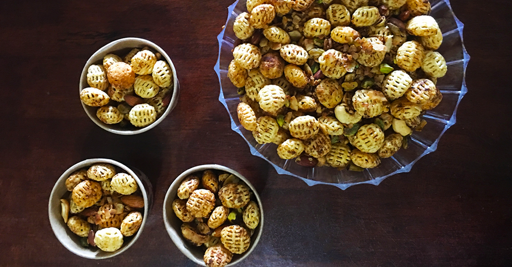 Large bowl and small cups filled with roasted, patterned snack bites mixed with nuts on a dark table.