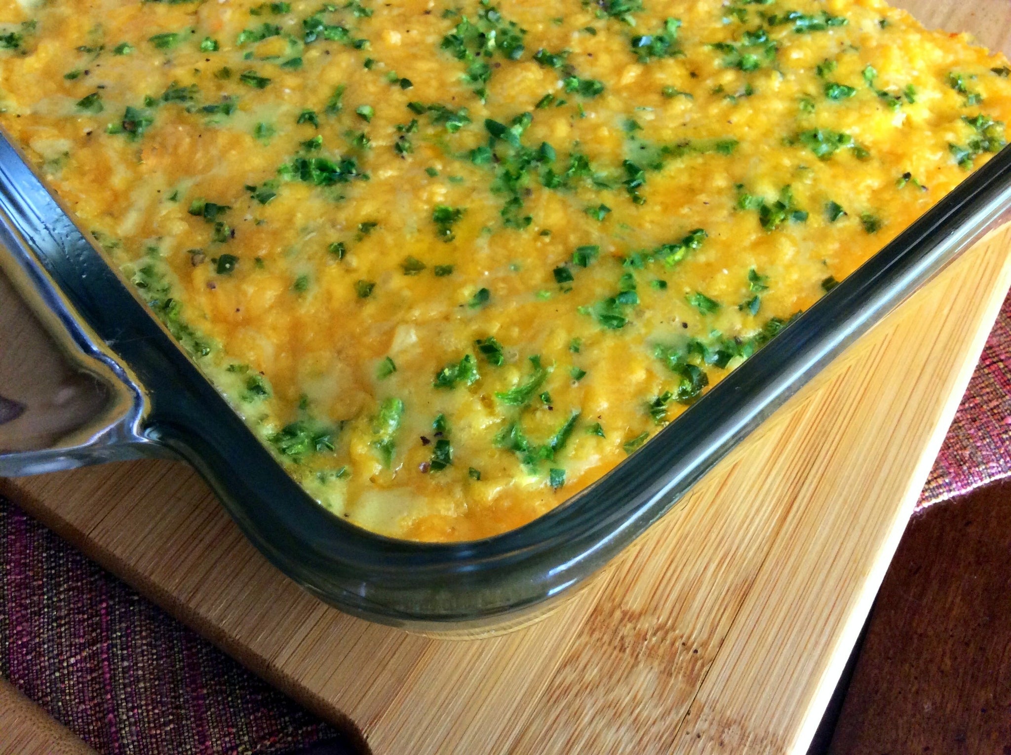 A baked casserole topped with melted cheese and chopped green herbs in a glass dish on a wooden board.