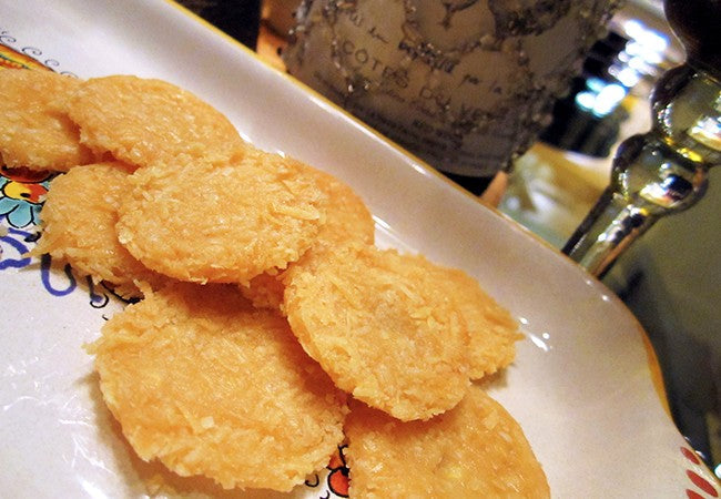 Round, crispy cheese wafers arranged on a decorative plate with a bottle and glass in the background.