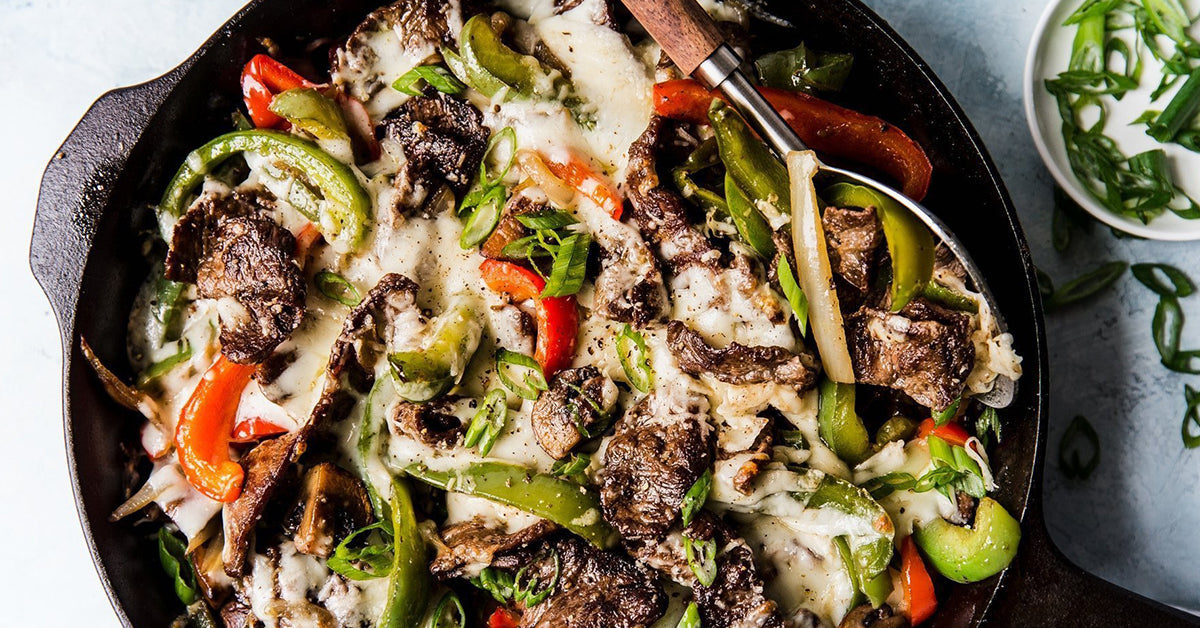 A cast iron skillet filled with sautéed beef, green and red bell peppers, and melted cheese, garnished with chopped green onions, sits on a light surface next to a small bowl of green onions.