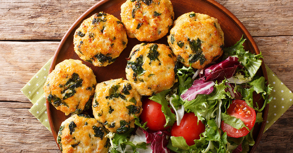 A wooden plate holds seven golden-brown, herb-studded patties alongside a fresh salad of mixed greens and sliced tomatoes, set on a rustic wooden table.