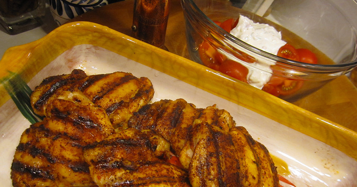 Grilled chicken thighs, seasoned and charred, are arranged on a decorative plate next to a bowl of cherry tomatoes and a creamy dip, set on a wooden table.