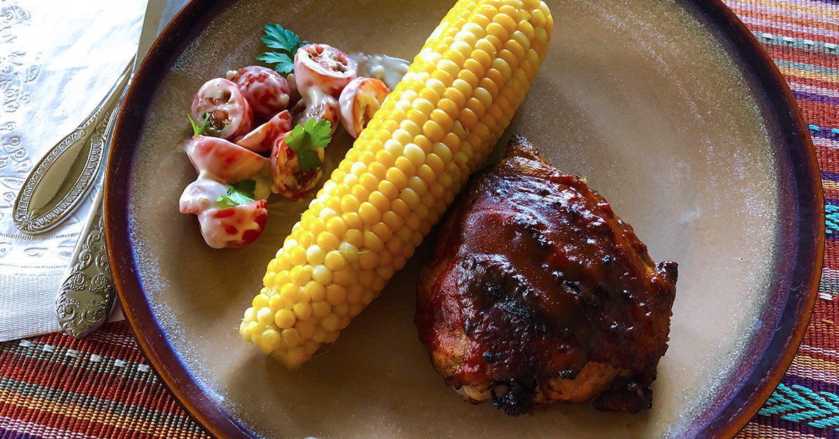 Plate with corn on the cob, barbecue chicken, and a small tomato salad with dressing.