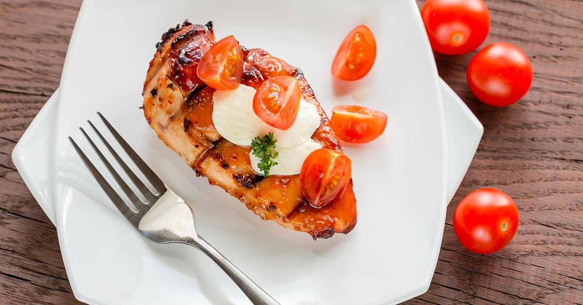 A grilled chicken breast topped with cherry tomatoes and a dollop of creamy sauce is served on a white plate, accompanied by a fork, with whole cherry tomatoes scattered on a wooden table.